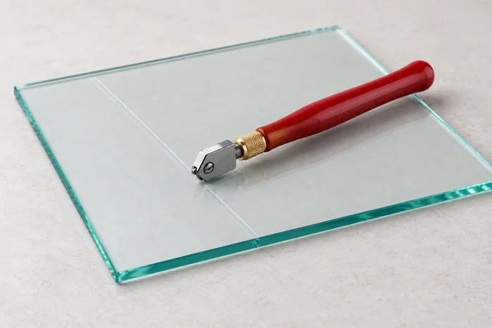 Handheld glass cutter resting on a clear glass sheet with a scored line